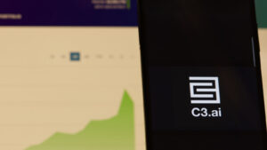 C3.ai (AI) のロゴがスマートフォンに表示され、コンピュータ画面が背景にグラフを表示し、AI 株を象徴している