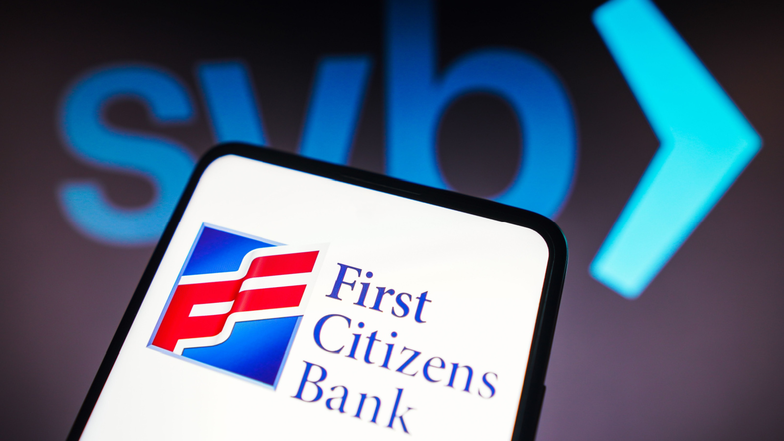 First Citizens Bank (FCNCA) logo displayed on smartphone in front of SVB logo in dark background behind phone