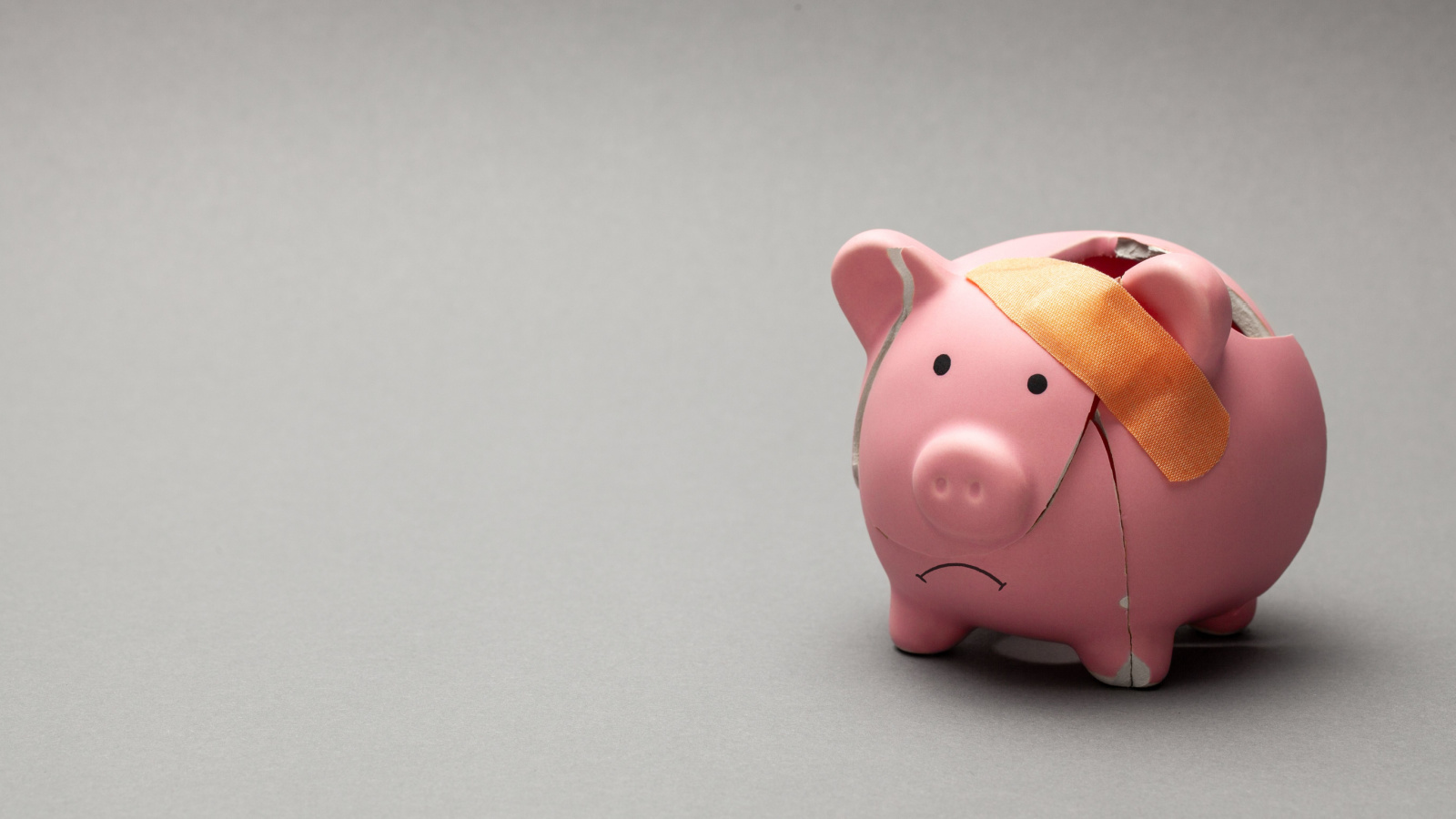 Photo of broken piggy bank with cracks on body and frown being held together by a band-aid. Gray background.