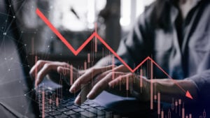 Grayish photo of investor's hands hovering over laptop with red stock graph showing downward arrow overlayed on top of the image