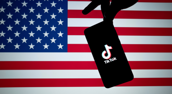 TikTok ban America. TikTok logo seen on the silhouette of smartphone hold in a hand with American flag on the back. US Banned Tik Tok concept. Best stocks to buy if TikTok is banned.