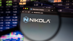 Nikola (NKLA) company logo on a website with blurry stock market developments in the background, seen on a computer screen through a magnifying glass.