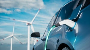 Photo of steel blue electric car being charged with wind silos and blue sky in the background