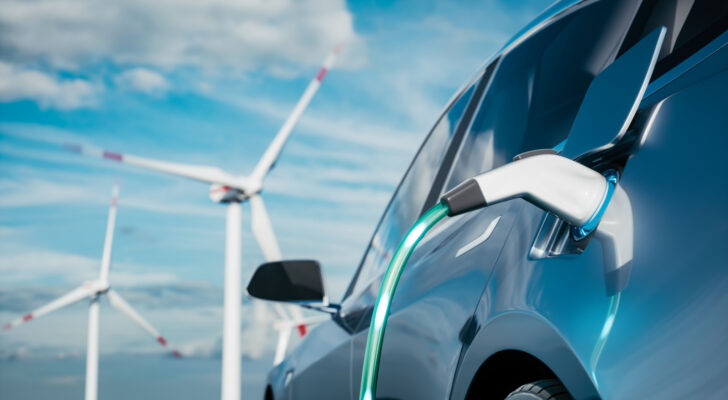 Photo of steel blue electric car being charged with wind silos and blue sky in the background. EV