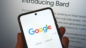 GoogleがBard AIを発表。 携帯電話の Google 検索バーと、背景にリリース情報が表示されます。 Google Bard AI 対 OpenAI ChatGPT。 GOOG株とGOOGL株。