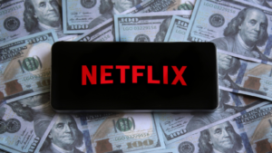 お金の山の上にスマートフォンに表示されたNetflix（NFLX）のロゴ。