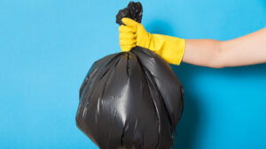 A hand with a yellow rubber glove holding up a big black trash bag in front of a teal background.
