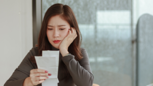 young woman look at pay bill holding ache at work. Tired woman employee looking at report on paper having headache. Stressful business deal concept. Gen Z and inflation