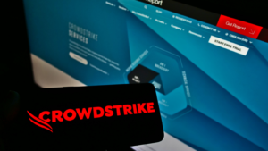 ウェブサイトの前の画面に、米国のソフトウェア会社 CrowdStrike Holdings Inc. (CRWD) のロゴが表示されたスマートフォンを持っている人物。スマートフォンのディスプレイに焦点を合わせている。加工なしの写真。