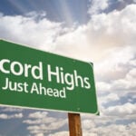 A green traffic sign that says Record Highs Just Ahead in front of a blue sky with white fluffy clouds. S&P 500 Stocks to Buy