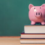 A pink piggy bank sitting on three stacked textbooks with a green chalkboard in the background.