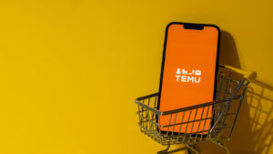 Smartphone displaying orange Temu logo in a miniature shopping cart against a yellow background