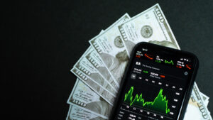 A pile of $100 bills on a black background with a smartphone displaying a stock chart on top.