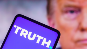 In this photo illustration, the Truth Social logo seen displayed on a smartphone with a photo of former US President Donald Trump displayed in the background. DJT stock