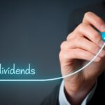 The word "dividends" written over a blue graph line being drawn by a business person's hand