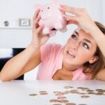 An image of a woman holding up a pink piggy bank with coins scattered on a flat surface below.