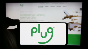 A person holding a mobile phone with the logo of Plug Power Inc., an American hydrogen cell company, in front of the company's website.  Focus on the phone display.  Unmodified photo.  Spare PLUG