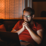 Portrait of attractive nerdy man with glasses is working late night on the computer in living room in home office. Meme stocks to sell