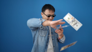 Young Chinese man wearing thug life glasses throwing dollars over isolated blue background. meme stocks