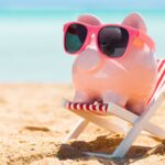 A pink piggy bank wearing pink sunglasses sitting in a beach chair in front of the ocean.