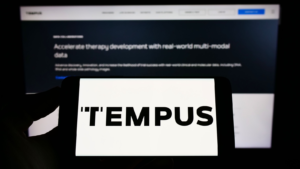 Person holding mobile phone with logo of American health care company Tempus Labs Inc. on screen in front of web page. Focus on phone display.