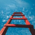 A red wooden ladder pointing up toward a blue sky with faint clouds.