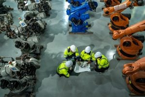 Four engineers, each wearing white hard hats and bright yellow and green jackets, squat around a rolled-out blueprint on a gray concrete floor. 